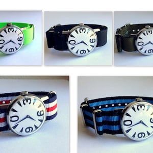May include: Five different wristwatches with white faces and black numbers. The watches have different colored nylon straps: green, black, camouflage, red and white striped, and blue and black striped.