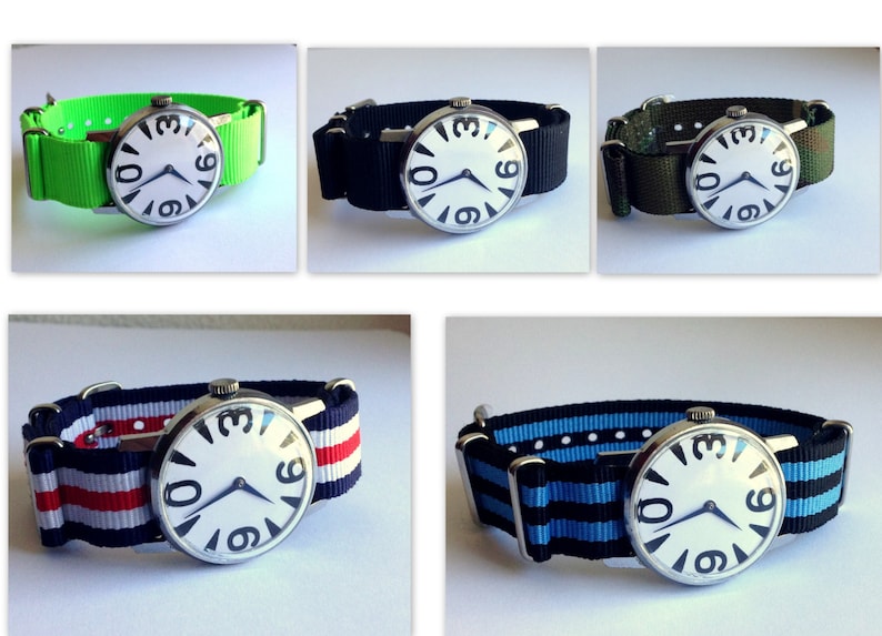 May include: Five different wristwatches with white faces and black numbers. The watches have different colored nylon straps: green, black, camouflage, red and white striped, and blue and black striped.