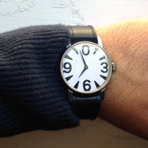 May include: A silver wristwatch with a white face and black numbers. The watch has a black leather band.