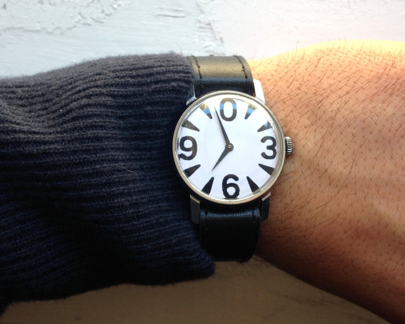 May include: A silver wristwatch with a white face and black numbers. The watch has a black leather band.