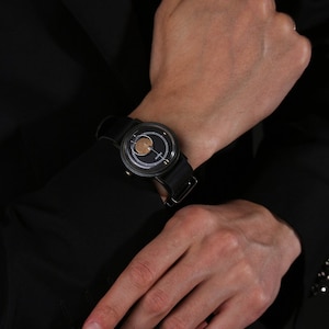 May include: A black wristwatch with a circular face and a black leather strap. The watch face has a gold-coloured centre and a dark background. The watch is worn on a wrist, with a black sleeve in the background.