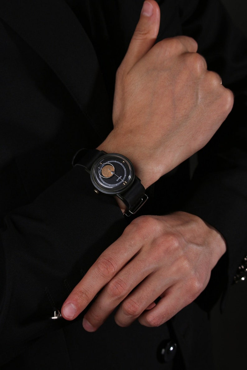 May include: A black wristwatch with a circular face and a black leather strap. The watch face has a gold-coloured centre and a dark background. The watch is worn on a wrist, with a black sleeve in the background.