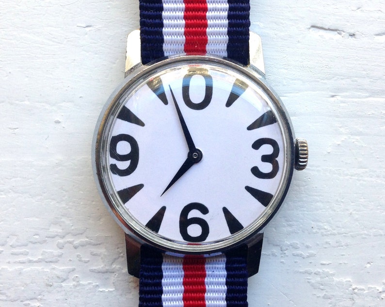 May include: A white wristwatch with black numbers and hands on a white face. The watch has a silver case and a red, white, and blue striped band.