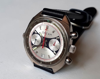 May include: A silver chronograph wristwatch with a white face and black accents. The watch has a black leather band and the words "OKEAH" and "23 камня" printed on the face.