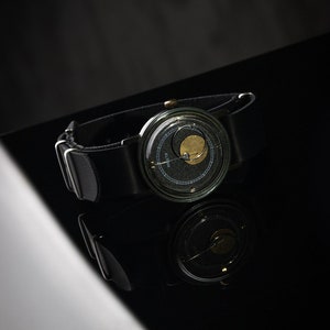 May include: A black wristwatch with a distinctive design. It has a dark face with a gold-coloured element and a black leather strap. The watch is set against a dark background, with its reflection visible.