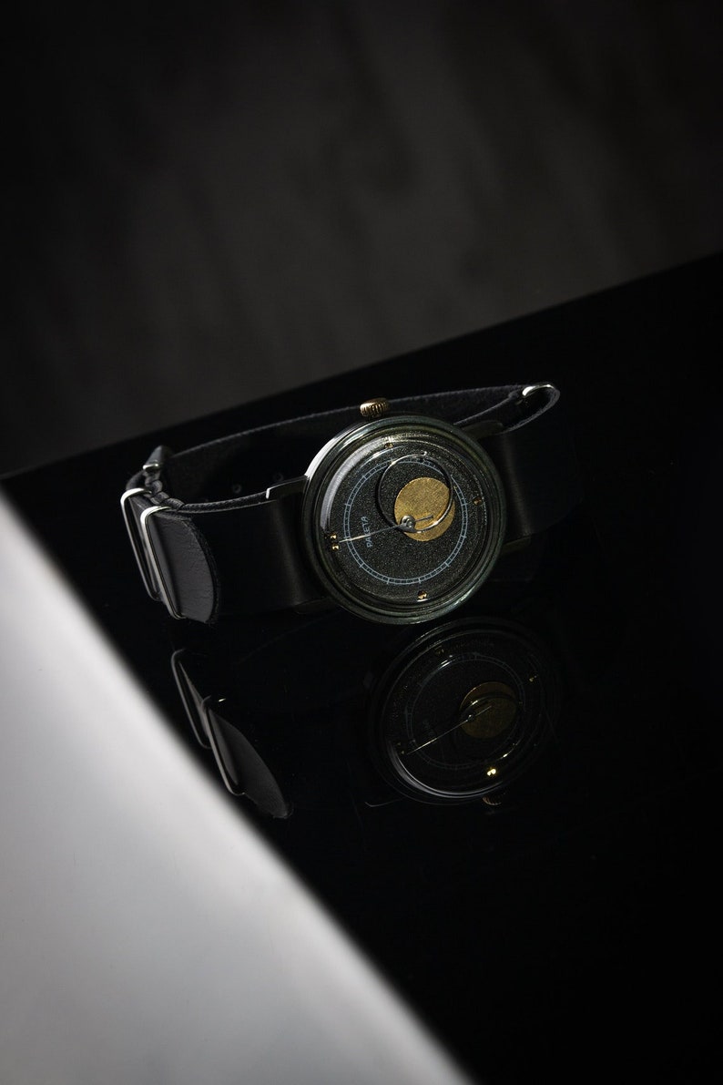 May include: A black wristwatch with a distinctive design. It has a dark face with a gold-coloured element and a black leather strap. The watch is set against a dark background, with its reflection visible.