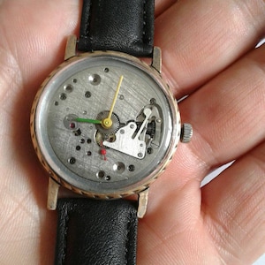 May include: A gold-tone wristwatch with a black leather band. The watch face is open, revealing the inner workings of the watch.