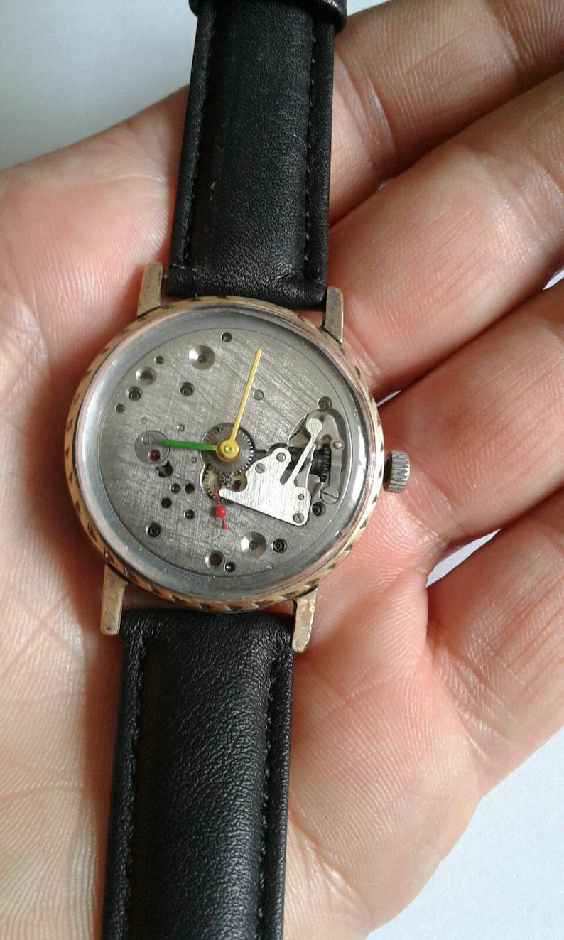 May include: A gold-tone wristwatch with a black leather band. The watch face is open, revealing the inner workings of the watch.