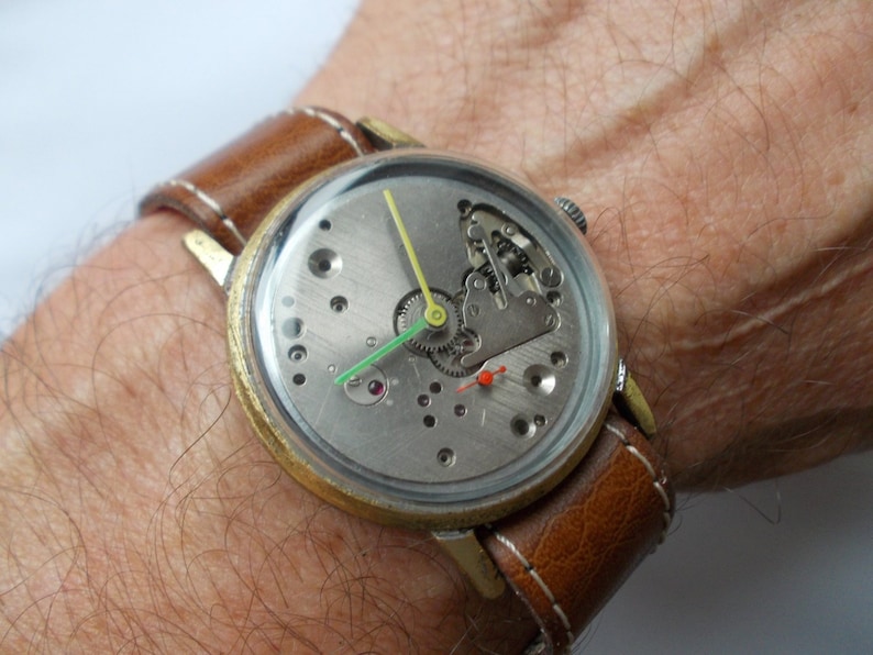 May include: A gold-tone wristwatch with a brown leather band. The watch face is clear and shows the inner workings of the watch mechanism.