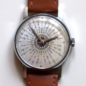 May include: A silver wristwatch with a brown leather band. The watch face has a compass design with the words "Nord", "Sud", "Ost", and "West" written around the compass.
