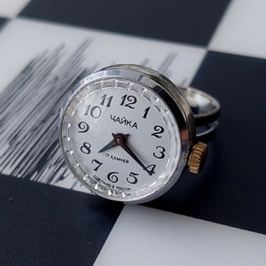 May include: A silver ring watch with a white face and black hands. The watch face has black numbers and the text "ЧАЙКА" and "17 КАМНЕЙ" in Cyrillic script. The watch is on a black and white chequered surface.