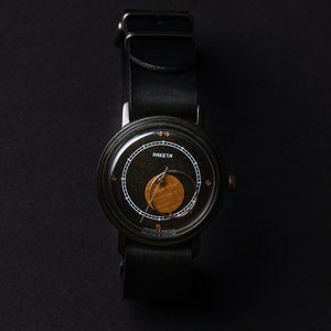 May include: A black wristwatch with a leather strap. The watch face is black with gold and white detailing, including the word "RAKETA". The watch has a round face and a black leather strap.