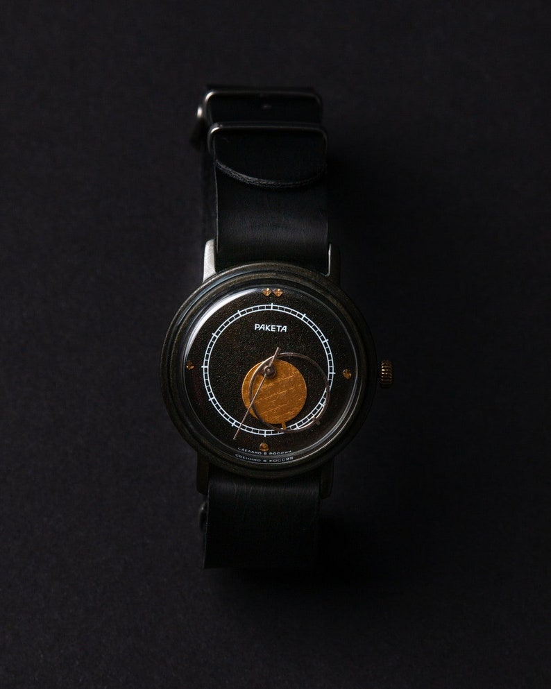 May include: A black wristwatch with a leather strap. The watch face is black with gold and white detailing, including the word "RAKETA". The watch has a round face and a black leather strap.