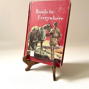 May include: A vintage book titled "Roads to Everywhere" with a red cover, featuring an illustration of a person with a rifle and a horse. The book is displayed on a small wooden easel.