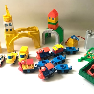 May include: A set of colorful plastic toy train tracks, cars, and buildings. The set includes a yellow bridge, a green tunnel, a gray building, a red and yellow tower, and a blue and red train engine with several cars.
