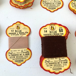 May include: Close-up of vintage nylon yarn spools in various colors. The spools have red and yellow labels with text that reads "Soft and Truly Durable" and "Spinnerin's Touchdown Yarn". The yarn colors include brown, tan, and green.
