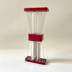 May include: A clear, hourglass-shaped kitchen timer with a red top, base, and center band. The timer contains a light-colored granular substance. The words "LIMIT" and "TIME" are printed on the red band.