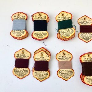 May include: A collection of vintage nylon reinforcing and mending yarn spools in various colors. Each spool has a decorative label with text that reads "Soft and Truly Durable" and "Touchdown Yarn".