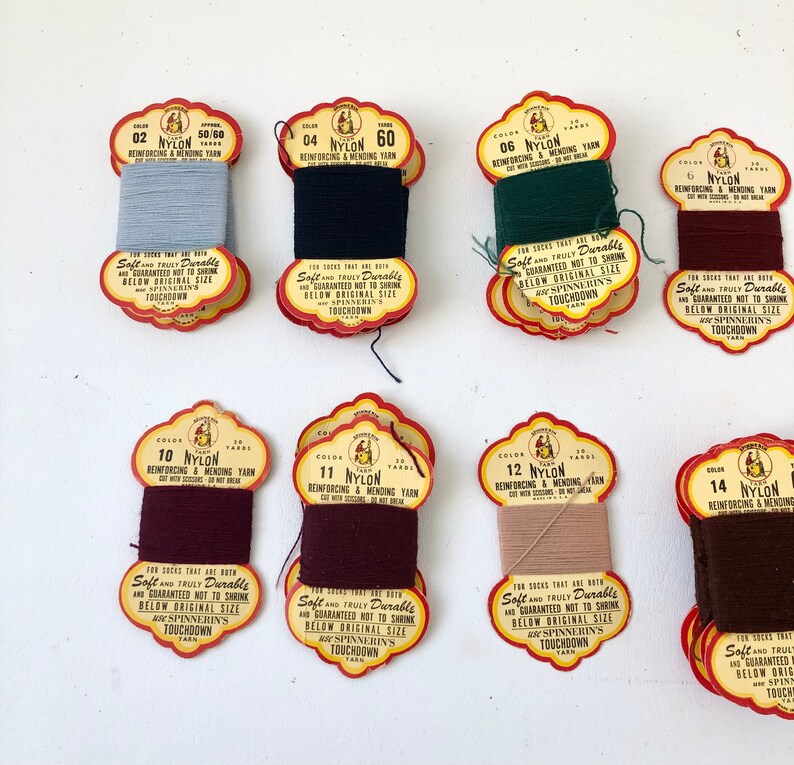 May include: A collection of vintage nylon reinforcing and mending yarn spools in various colors. Each spool has a decorative label with text that reads "Soft and Truly Durable" and "Touchdown Yarn".
