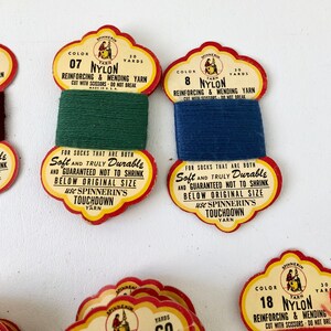May include: Close-up of vintage nylon yarn spools in green and blue. Each spool has a decorative label with text that reads "Reinforcing & Mending Yarn" and "Touchdown Yarn". The labels are red, yellow, and white.