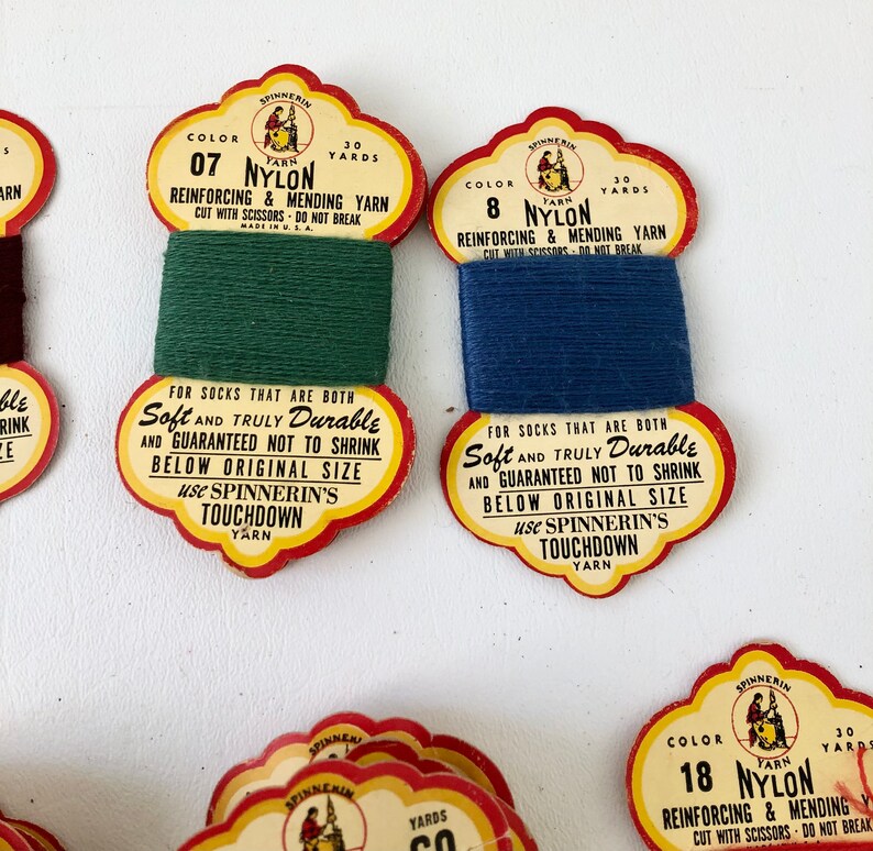 May include: Close-up of vintage nylon yarn spools in green and blue. Each spool has a decorative label with text that reads "Reinforcing & Mending Yarn" and "Touchdown Yarn". The labels are red, yellow, and white.