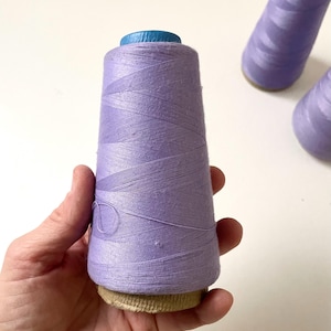 May include: A hand holds a large spool of lavender thread. The thread is tightly wound around a cardboard core, topped with a blue plastic cap. Two additional spools of thread are visible in the background, suggesting a crafting or sewing theme.