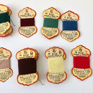 May include: A collection of eight spools of nylon yarn in various colors, including green, burgundy, teal, blue, tan, brown, yellow, and red. Each spool has a vintage label with text.
