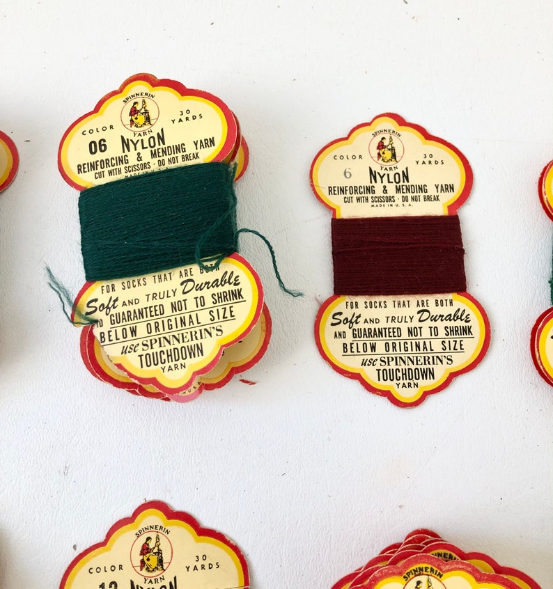 May include: Close-up of several vintage spools of nylon yarn. Each spool has a colorful label with text that reads "Reinforcing & Mending Yarn" and "Soft and Truly Durable". The yarn colors include green and burgundy.