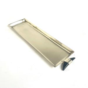 Peut inclure: Un plateau rectangulaire en métal argenté avec une finition polie et une surface lisse. Le plateau présente une poignée à une extrémité, ornée d'un élément décoratif sombre, incurvé et en forme de corne, fixé par un support métallique.