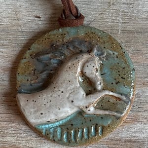 May include: A round ceramic pendant with a horse design. The pendant features a white horse with a brown leather cord. The horse is set against a teal and brown background. The pendant is a unique piece of jewelry.