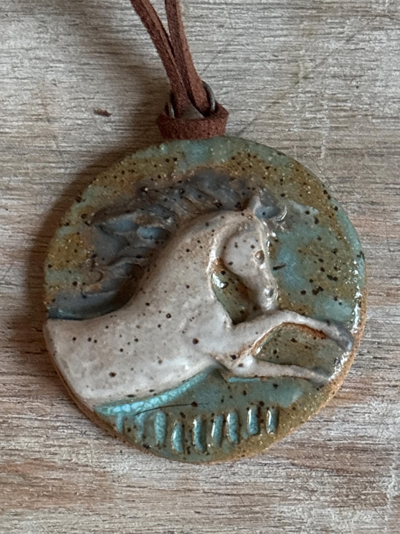 May include: A round ceramic pendant with a horse design. The pendant features a white horse with a brown leather cord. The horse is set against a teal and brown background. The pendant is a unique piece of jewelry.