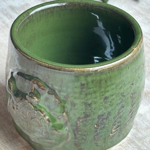 May include: A handcrafted ceramic mug with a glossy green glaze and a textured, organic design. The interior is a darker green, and the rim has a subtle gold accent. The mug has a unique, earthy aesthetic.