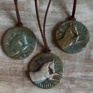 May include: Three ceramic horse pendant necklaces. Each pendant is a circular disc with a raised horse design. The pendants are in shades of blue, brown, and white, with brown leather cords. The necklaces are displayed on a light wood surface.