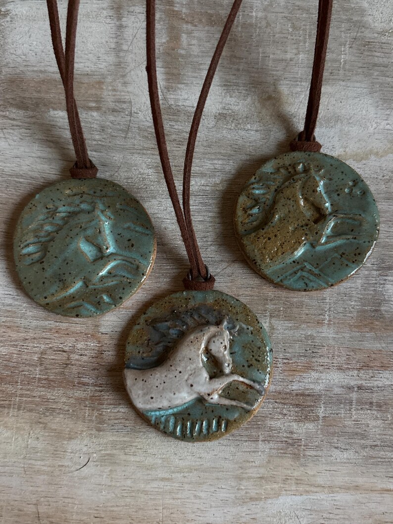May include: Three ceramic horse pendant necklaces. Each pendant is a circular disc with a raised horse design. The pendants are in shades of blue, brown, and white, with brown leather cords. The necklaces are displayed on a light wood surface.