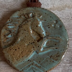 May include: A round, ceramic pendant with a horse head design. The pendant is a speckled teal and brown color, with a brown leather cord attached at the top. The horse head is raised and detailed.