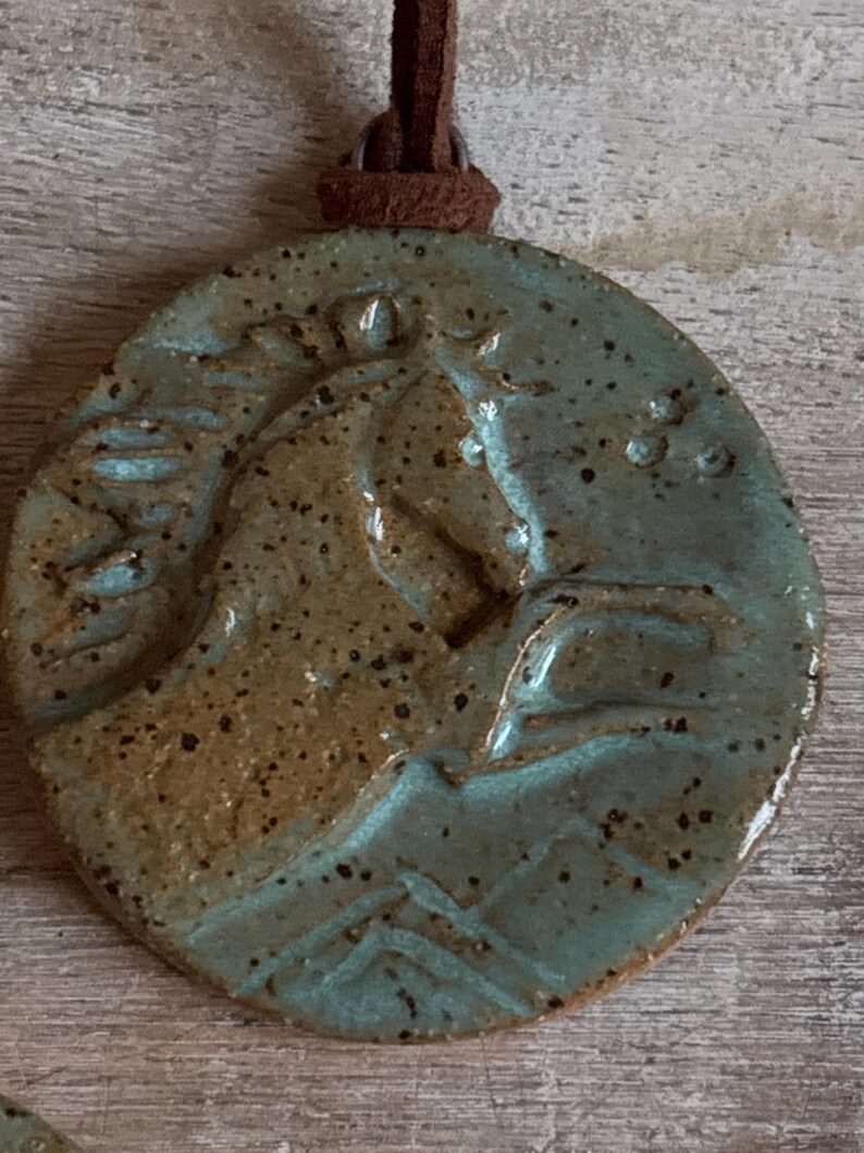 May include: A round, ceramic pendant with a horse head design. The pendant is a speckled teal and brown color, with a brown leather cord attached at the top. The horse head is raised and detailed.