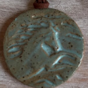 May include: A round, ceramic pendant with a horse's head design in relief. The pendant is a mottled teal color with a brown leather cord for wearing. The pendant is approximately 2 inches in diameter.