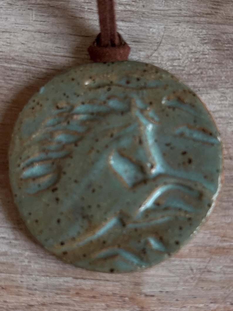 May include: A round, ceramic pendant with a horse's head design in relief. The pendant is a mottled teal color with a brown leather cord for wearing. The pendant is approximately 2 inches in diameter.