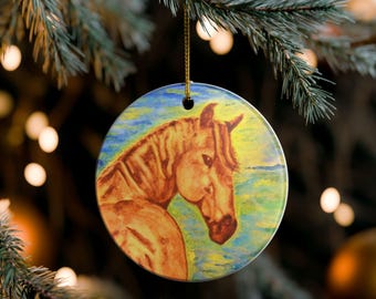 Könnte beinhalten: Runder Weihnachtsbaumschmuck aus Keramik mit einem Aquarellgemälde eines Pferdekopfes. Das Pferd ist in Braun- und Beigetönen vor einem Hintergrund aus Blau, Gelb und Grün dargestellt. Der Schmuck hängt an einem goldenen Faden.