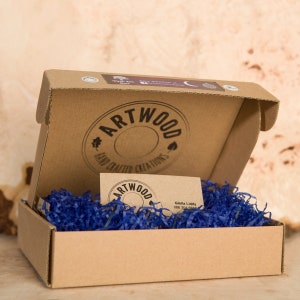 May include: A brown cardboard box with the "ARTWOOD HAND CRAFTED CREATIONS" logo on the lid and a business card inside. The box is filled with blue shredded paper. The business card displays the "ARTWOOD" logo and contact details.