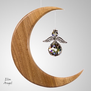 May include: A wooden crescent moon with a crystal angel ornament suspended from a string. The angel features silver wings and a faceted crystal sphere. The moon is crafted from light brown wood, with the text "Elm Angel" in the lower left.