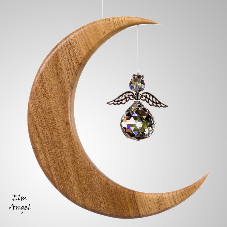 May include: A wooden crescent moon with a crystal angel ornament suspended from a string. The angel features silver wings and a faceted crystal sphere. The moon is crafted from light brown wood, with the text "Elm Angel" in the lower left.