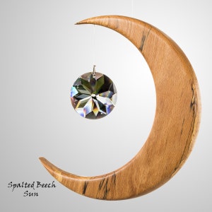 May include: A wooden crescent moon with a clear, faceted crystal sun suspended from a thin wire. The moon is made of spalted beech wood, with dark streaks. The text "Spalted Beech Sun" is at the bottom left.