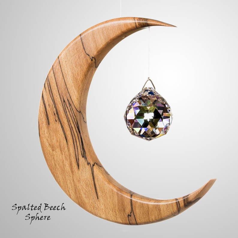 May include: A wooden crescent moon sculpture with a hanging crystal sphere. The moon is made of spalted beech wood, with dark streaks. The crystal sphere reflects multiple colours. Text on the bottom left reads "Spalted Beech Sphere."
