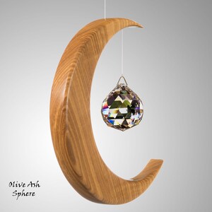 May include: A wooden crescent-shaped sculpture with a hanging crystal sphere. The sculpture is made of light brown wood and has a smooth finish. The crystal sphere is faceted and reflects multiple colours. The text "Olive Ash Sphere" is at the bottom.