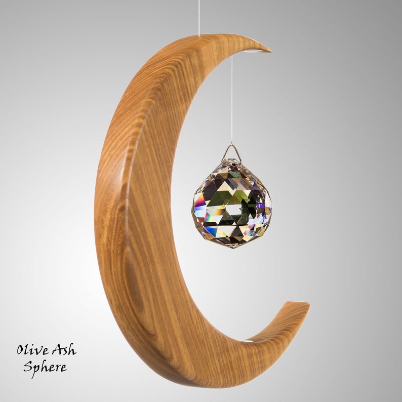 May include: A wooden crescent-shaped sculpture with a hanging crystal sphere. The sculpture is made of light brown wood and has a smooth finish. The crystal sphere is faceted and reflects multiple colours. The text "Olive Ash Sphere" is at the bottom.