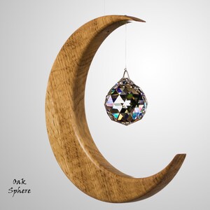 May include: A wooden crescent moon sculpture with a hanging faceted crystal sphere. The sculpture is made of light brown wood. The text "Oak Sphere" is in the lower left corner. The crystal sphere hangs in the center.