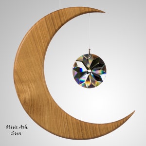 May include: A wooden crescent moon with a clear, faceted crystal sun suspended from a string. The moon is made of light brown wood. The text "Olive Ash Sun" is at the bottom left.