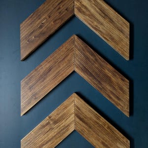 May include: Three chevron-shaped wooden wall art pieces arranged vertically. Each piece is crafted from wood with a dark brown stained finish, showcasing a visible natural wood grain pattern and sharp, angled mitered joints at the center point.