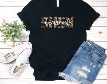 Könnte beinhalten: Schwarzes T-Shirt mit dem Text "SHSU bearkats", wobei das Wort "bearkats" im Leopardenmuster gehalten ist. Das Shirt wird mit Jeansshorts und weißen Turnschuhen auf einer Holzoberfläche präsentiert.